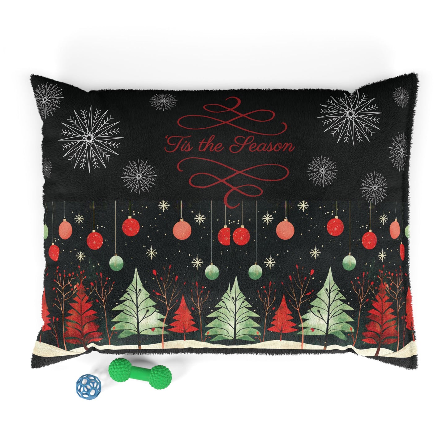 A rectangular black throw pillow with a festive Christmas design, featuring red and green ornaments, evergreen trees, snowflakes, and the text 'Tis the Season' in red script. The pillow is shown on a white background with two small dog toys in front.