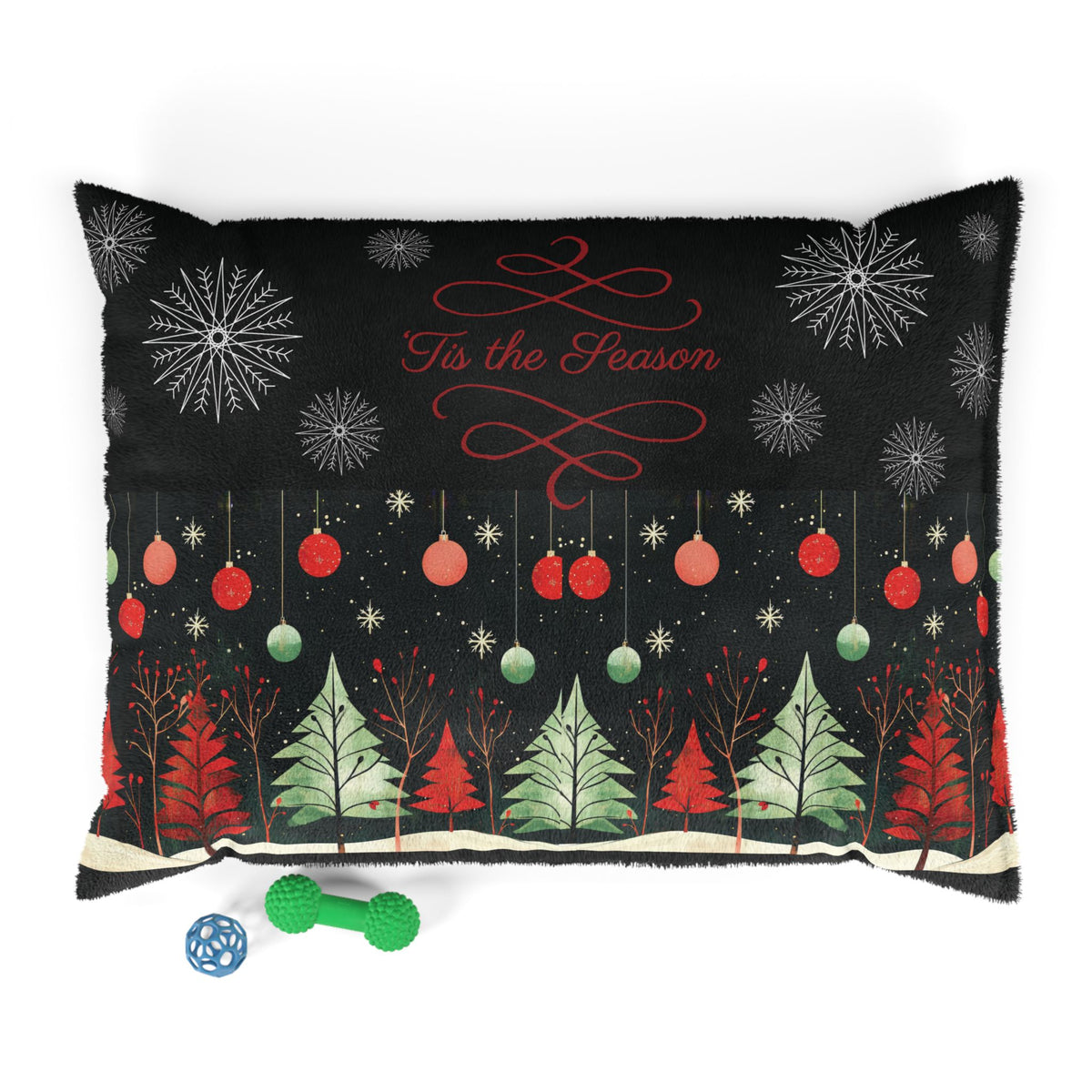 A rectangular black throw pillow with a festive Christmas design, featuring red and green ornaments, evergreen trees, snowflakes, and the text 'Tis the Season' in red script. The pillow is shown on a white background with two small dog toys in front.