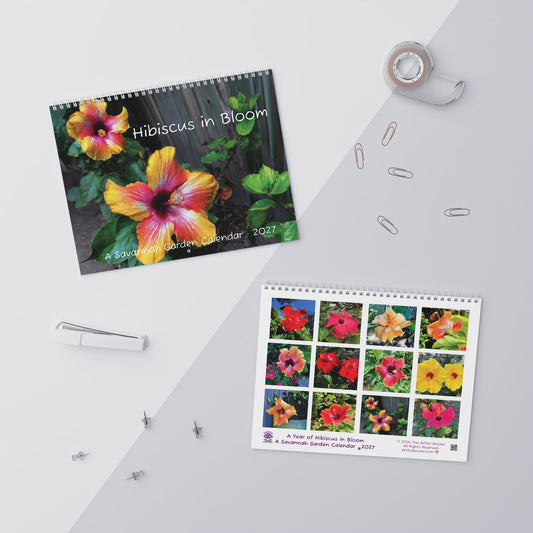 Two spiral-bound Hibiscus in Bloom garden calendars are displayed on a light gray surface with decorative stationery props including tape, paper clips, a pen, and pushpins. One calendar shows a large hibiscus flower cover, and the other shows a 3x4 grid of colorful hibiscus flowers.